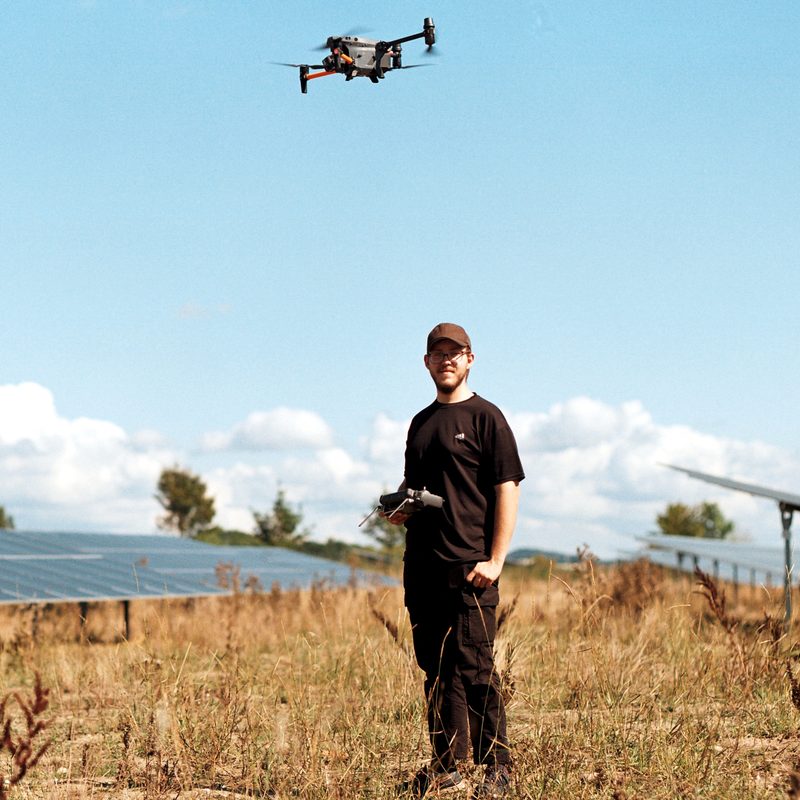 Florian Wagner bei der Arbeit im Feld: mit der Fernsteuerung vor einer Freiflächen-Photovoltaikanlage, Drohne im Flug